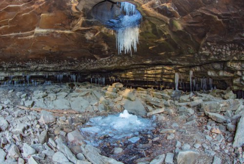 Icicles hang from a rocky cave ceiling above a frozen pool and scattered stones on the cave floor.
