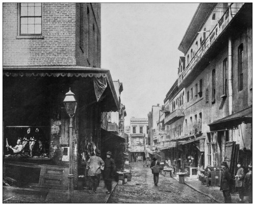 Historic street scene with buildings, shops, and people walking in a bustling urban environment.