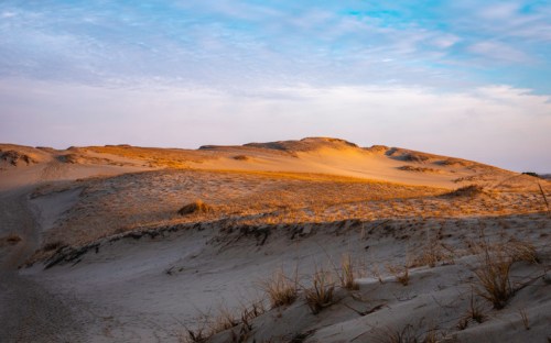 A serene landscape of sand dunes under a blue sky, with soft golden light illuminating the sandy terrain.