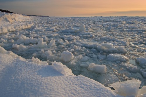 A frozen landscape with ice chunks scattered across a body of water, under a soft sunset sky.