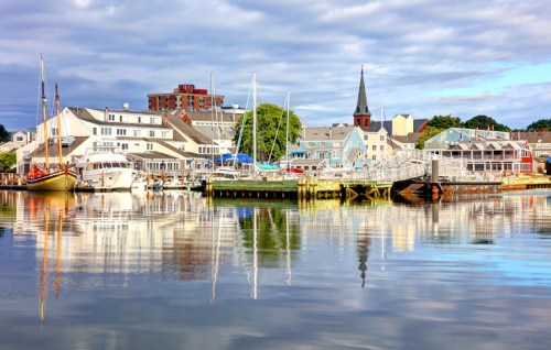 A serene harbor scene with boats, colorful buildings, and a church steeple reflected in calm water under a cloudy sky.