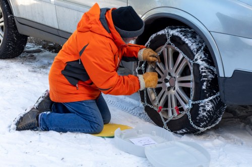 A person in an orange jacket is kneeling in snow, installing tire chains on a vehicle's wheel.