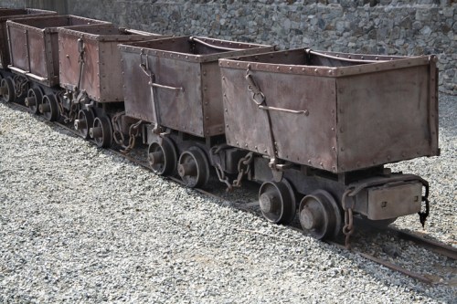 A row of vintage mining carts on tracks, resting on a gravel surface.