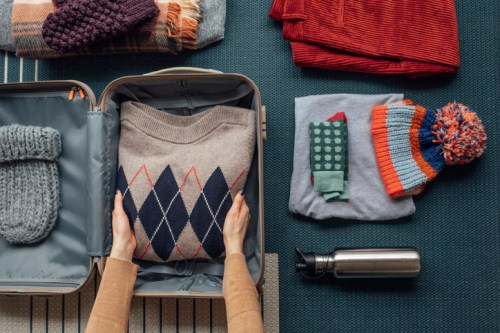 A person packs a suitcase with folded sweaters, hats, and a water bottle on a patterned surface.