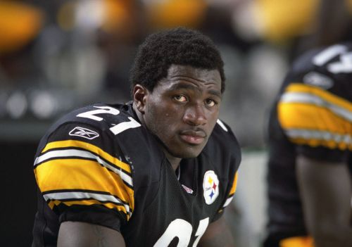 A football player in a black and gold jersey sits on the sidelines, looking thoughtfully into the distance.
