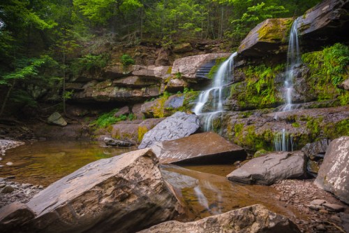 A serene waterfall cascades over moss-covered rocks into a calm pool, surrounded by lush green trees.
