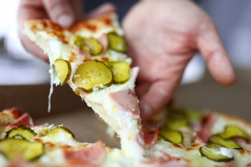 A hand holding a slice of pizza topped with pickles and ham, with melted cheese stretching from the pie.
