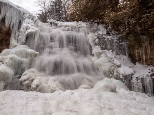 A frozen waterfall cascading over icy rocks, surrounded by snow and frosty trees.
