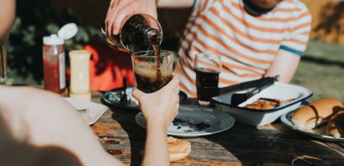 A person pours a drink into a glass at a casual outdoor gathering with food on the table.