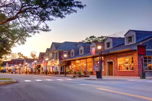 Charming street lined with shops and cafes, illuminated by warm lights at sunset. American flags adorn the buildings.