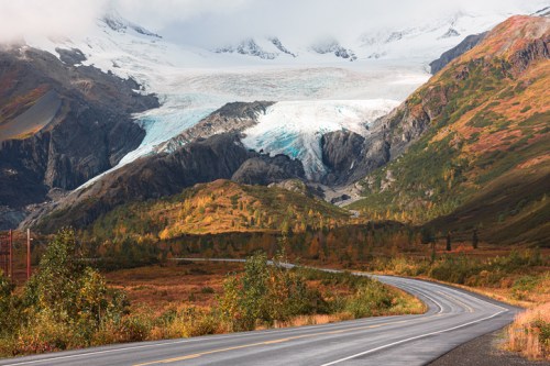 A winding road leads through colorful autumn foliage, with a glacier and mountains in the background under a cloudy sky.