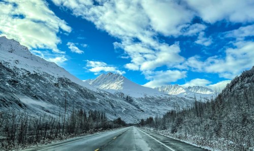 A snowy mountain landscape with a clear blue sky, viewed from an empty road stretching into the distance.