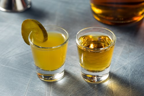 Two shot glasses on a metal surface: one with a yellow drink and a pickle slice, the other with a golden liquid.