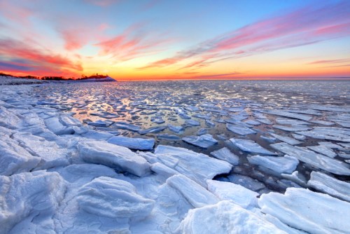 A serene winter landscape with ice formations on water, reflecting a colorful sunset sky with pink and orange hues.