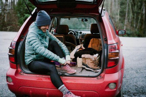 A person sits in the back of a red car, putting on hiking boots, surrounded by outdoor gear and a forest backdrop.