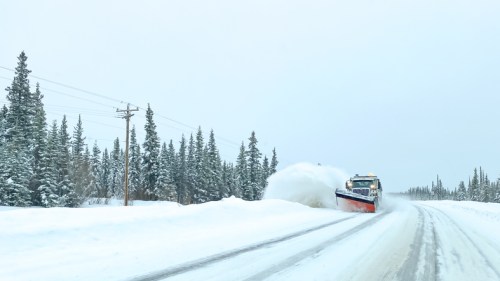 A snowplow clears a snowy road surrounded by tall evergreen trees under a gray sky.