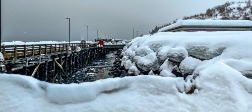 A snowy pier extends into a frozen body of water, surrounded by snow-covered rocks and a cloudy sky.