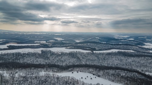 Aerial view of a snowy landscape with rolling hills and overcast skies, featuring patches of trees and open fields.