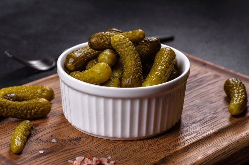 A white bowl filled with pickles, placed on a wooden cutting board, with a few pickles scattered around.