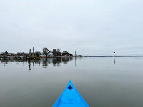 A blue kayak's bow facing a calm, reflective lake with a cloudy sky and distant shoreline.