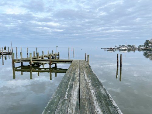 A calm waterfront scene featuring a wooden dock extending into still water, with cloudy skies above.