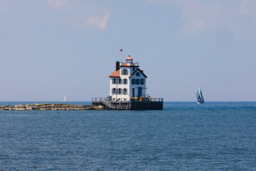 A white lighthouse with a red roof stands on a pier, with sailboats in the background on a calm blue lake.