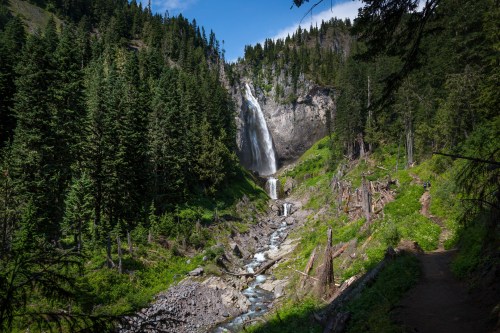 A scenic waterfall cascades down rocky cliffs, surrounded by lush green trees and a winding stream below.