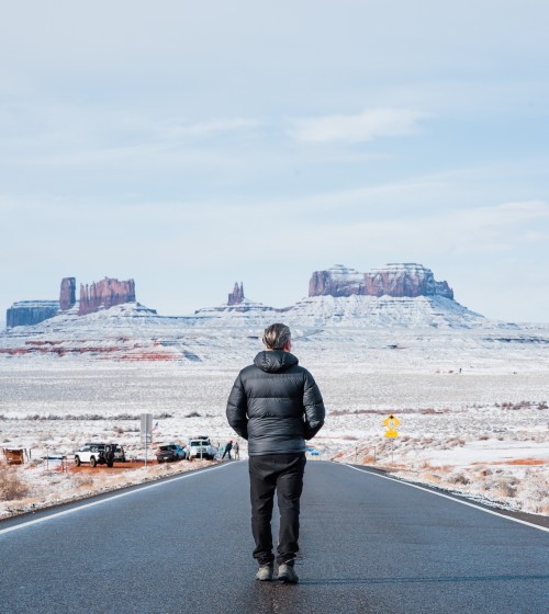 A person in a black puffer jacket stands on a snowy road, facing distant rock formations under a clear blue sky.