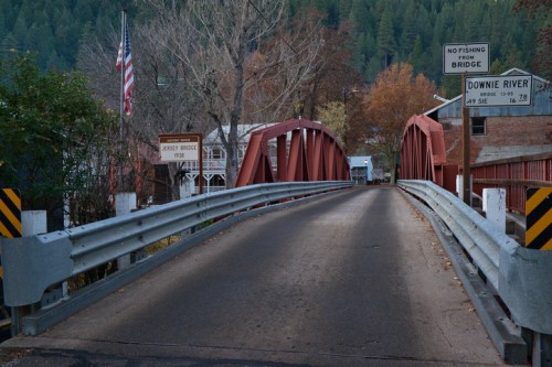 A red metal bridge spans a road with signs for fishing restrictions and directions to the Downie River. Trees in autumn colors surround it.