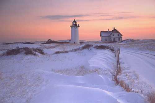 A snowy landscape at sunset featuring a lighthouse and a nearby house, with soft pastel colors in the sky.