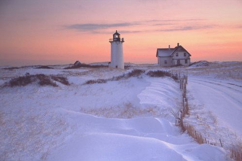 A snowy landscape at sunset featuring a lighthouse and a nearby house, with soft pastel colors in the sky.