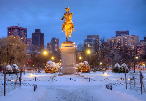 A golden statue of a mounted figure stands in a snowy park, with a city skyline illuminated in the background at dusk.