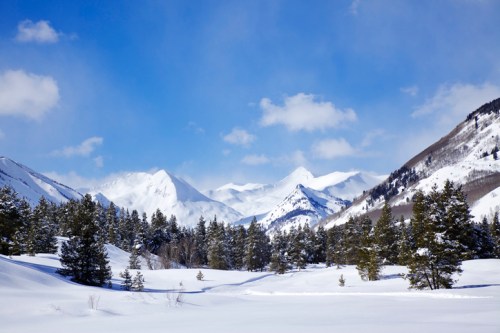 Snow-covered mountains under a blue sky, surrounded by evergreen trees and a serene winter landscape.