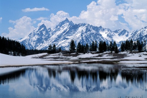 Snow-capped mountains reflect in a calm lake, surrounded by evergreen trees and a clear blue sky with fluffy clouds.