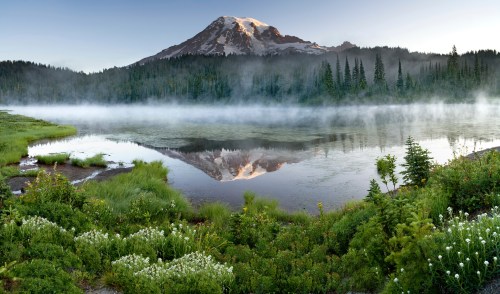 A serene lake reflects a snow-capped mountain, surrounded by lush greenery and mist in the early morning light.
