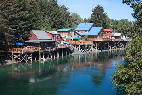 Colorful houses on stilts along a calm river, surrounded by lush trees and clear blue skies.