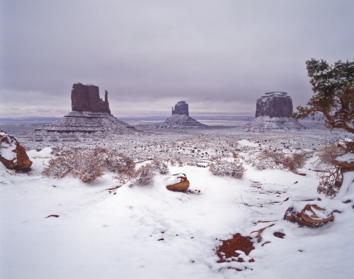 Snow-covered landscape featuring three iconic rock formations under a cloudy sky. Sparse vegetation in the foreground.