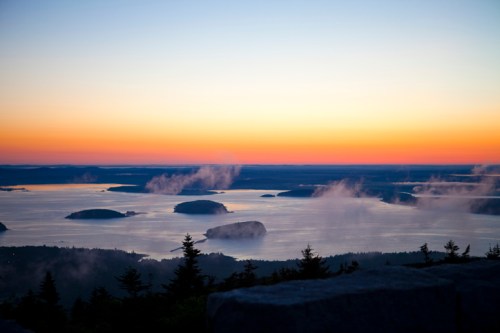 A serene sunset over a calm bay, with islands and misty clouds reflecting vibrant colors in the sky.
