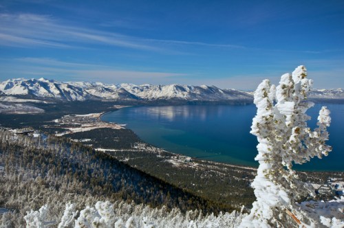 A snowy landscape overlooking a lake surrounded by mountains under a clear blue sky.