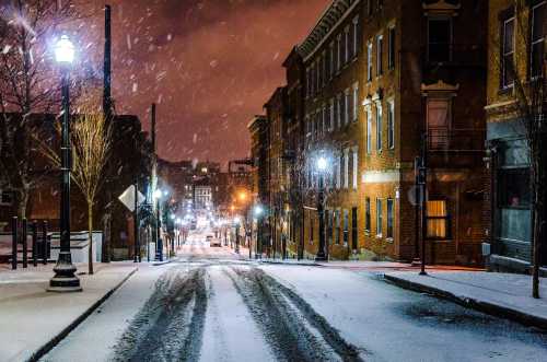 A snowy street at night, lined with brick buildings and streetlights, creating a serene winter scene.