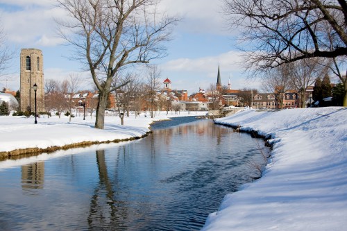 A serene winter scene featuring a snow-covered landscape, a calm river, and a distant town with church spires.