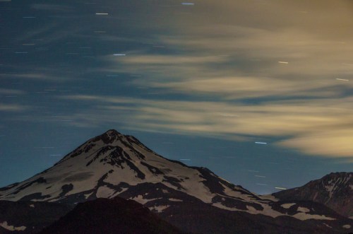 A snow-capped mountain under a starry sky with streaks of clouds and stars, creating a serene nighttime landscape.