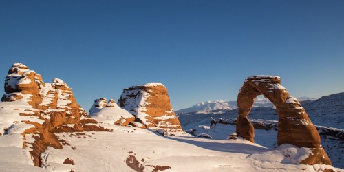 Snow-covered landscape featuring Delicate Arch and surrounding rock formations under a clear blue sky.