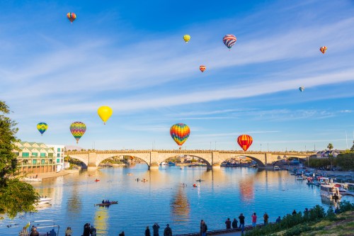 Colorful hot air balloons float over a river with a bridge, reflecting in the water under a clear blue sky.