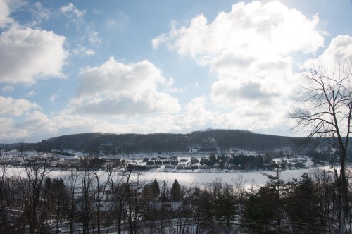 A snowy landscape with a frozen lake, hills in the background, and a cloudy blue sky.