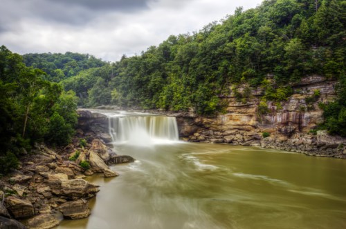 A serene waterfall cascades into a calm river, surrounded by lush green trees and rocky cliffs under a cloudy sky.
