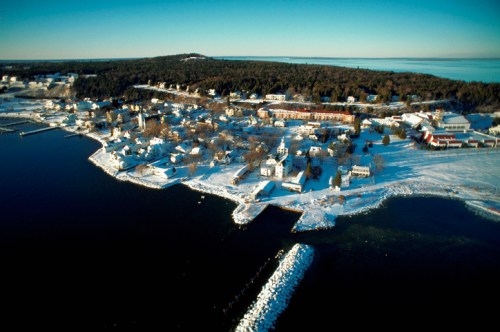 Aerial view of a snowy coastal town with houses, trees, and a calm blue sea in the background.