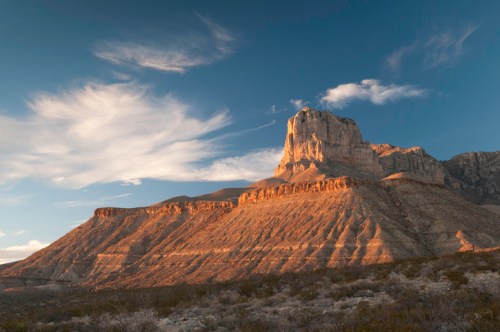A dramatic mountain landscape with layered rock formations under a blue sky with wispy clouds.