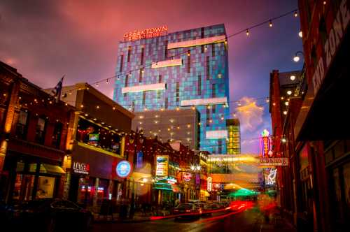 A vibrant city street at dusk, featuring colorful lights, a modern hotel, and lively storefronts.