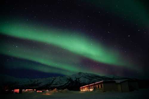 A stunning display of the Northern Lights illuminates the night sky over snow-covered mountains and a building.